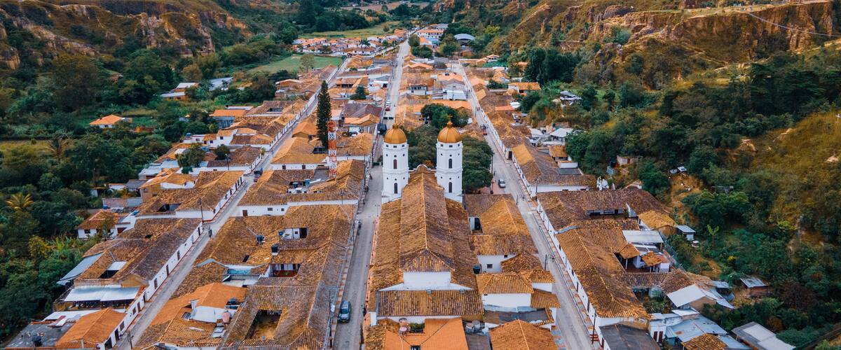 An aerial view of a town with terracotta-roofed buildings, a prominent church with two towers, and surrounding mountainous terrain.