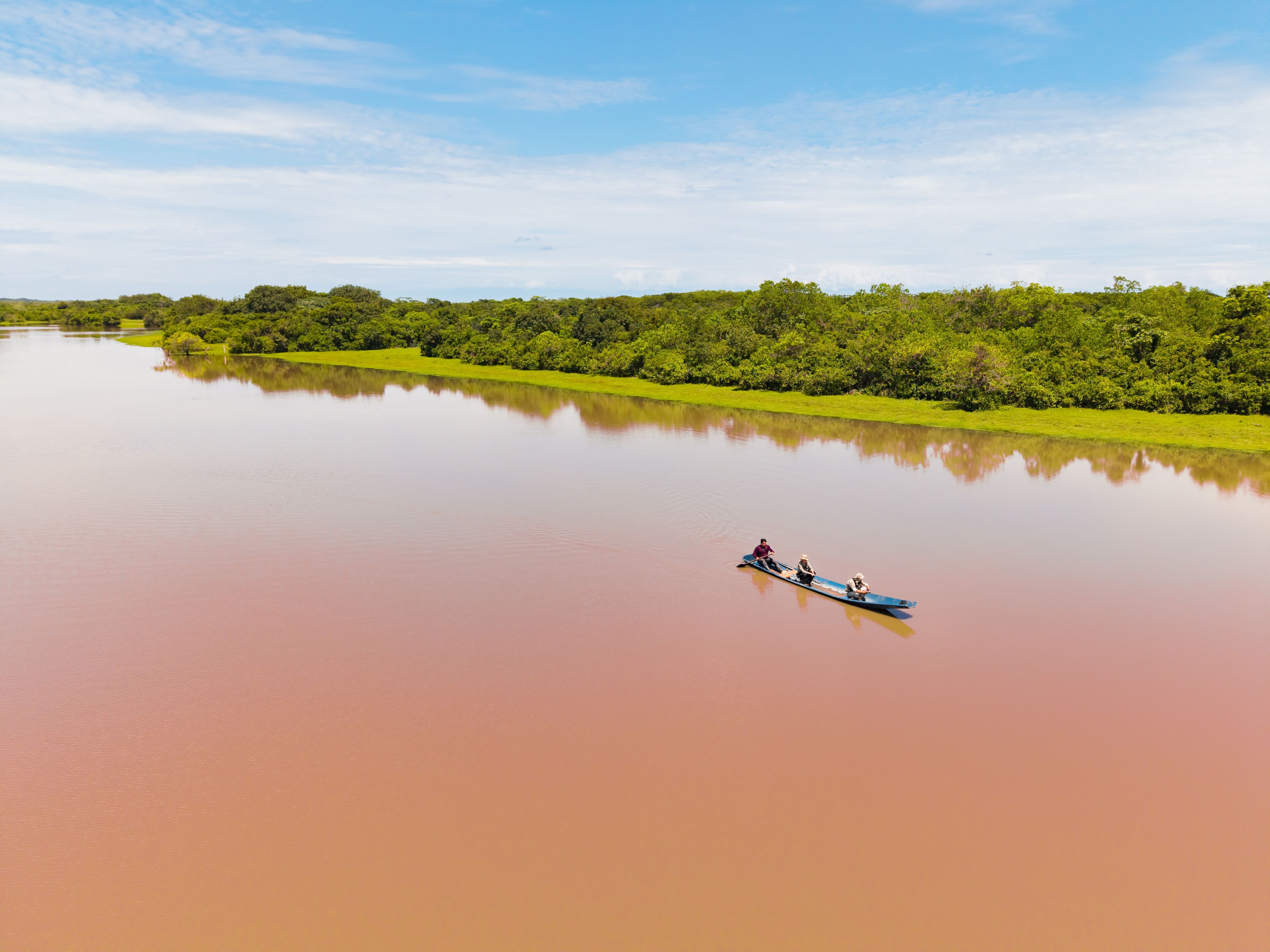 Aerial view of a boat sailing along the Guanapalo River, its waters reflecting the sky under the bright sun, framed by lush green trees, San Luis de Palenque, Casanare, Colombia.