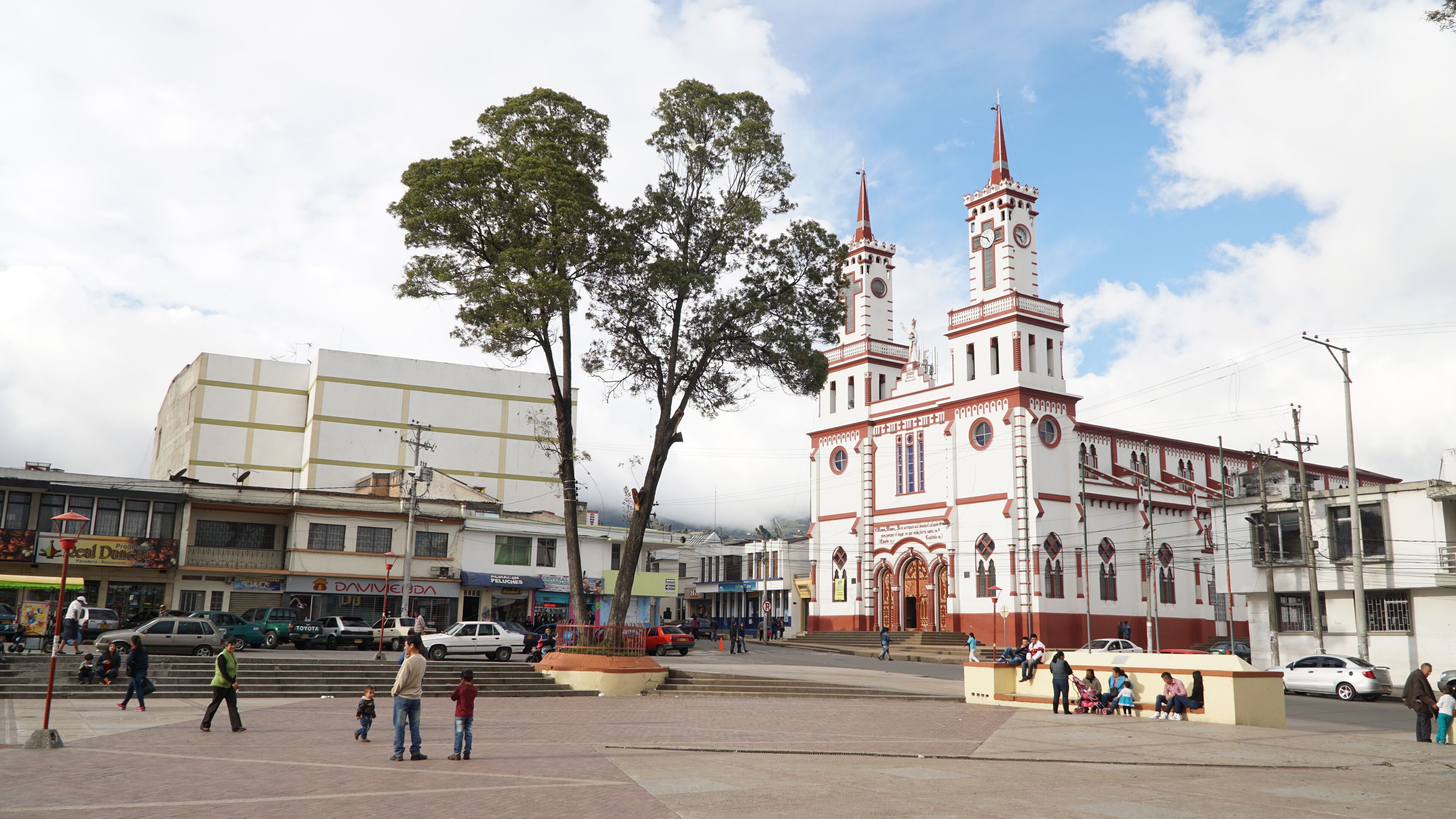 A plaza with a prominent church featuring a tall spire and clock tower, surrounded by trees and various buildings.