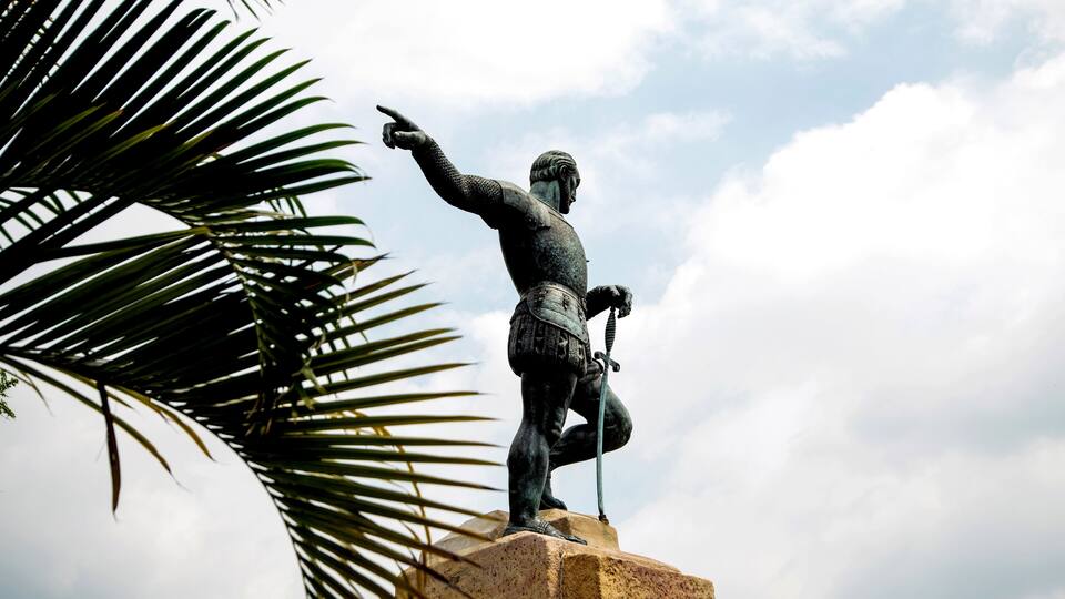 A statue of a historical figure on a pedestal, with a palm tree in the foreground.