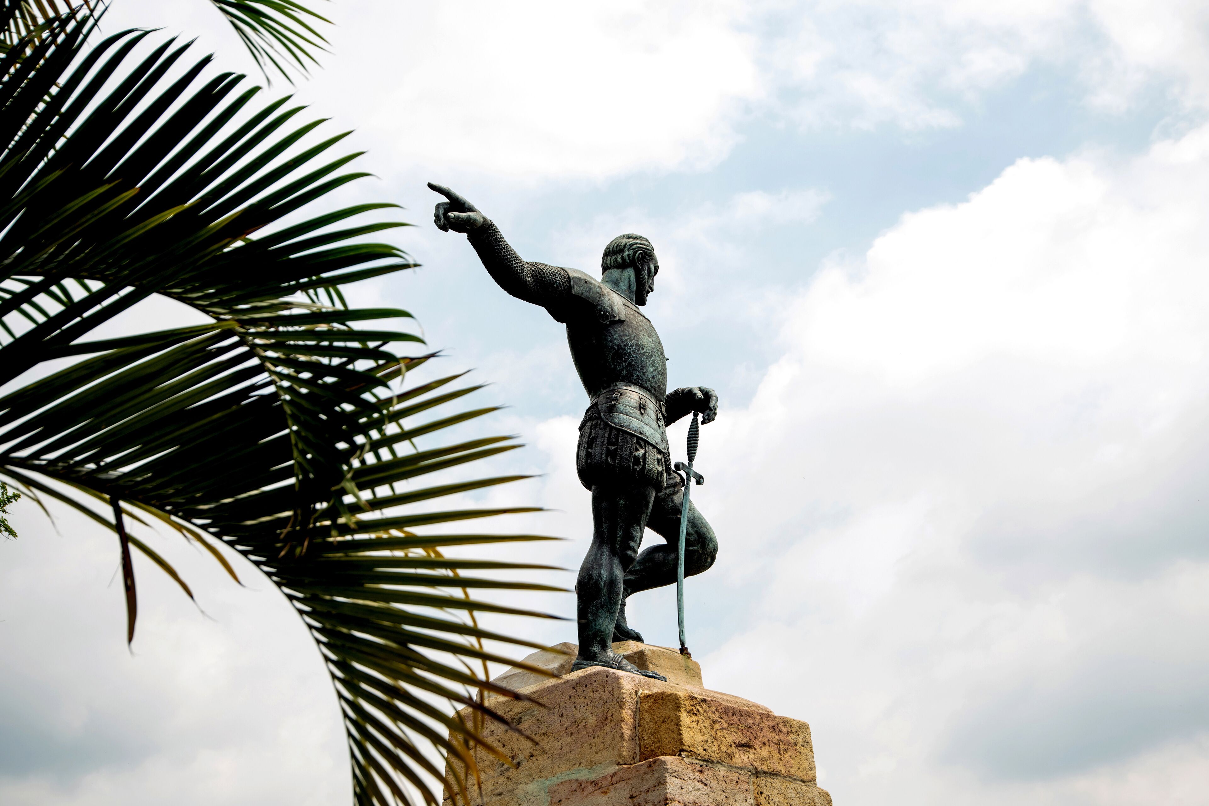 A statue of a historical figure on a pedestal, with a palm tree in the foreground.