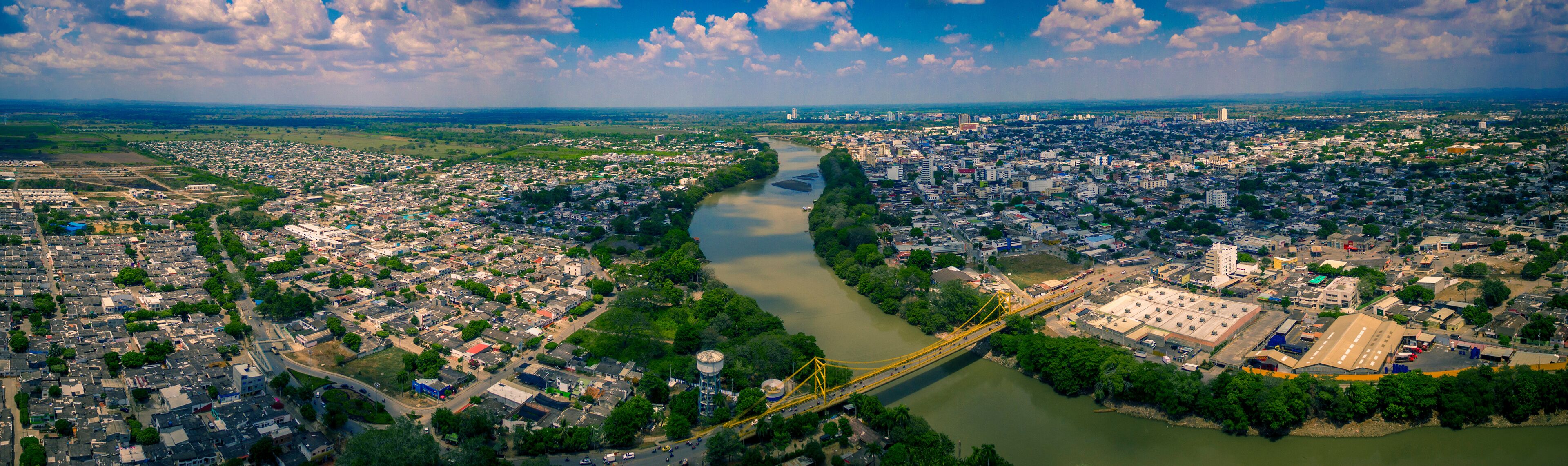 An aerial view of a city with a river running through it, a bridge, and surrounding greenery.