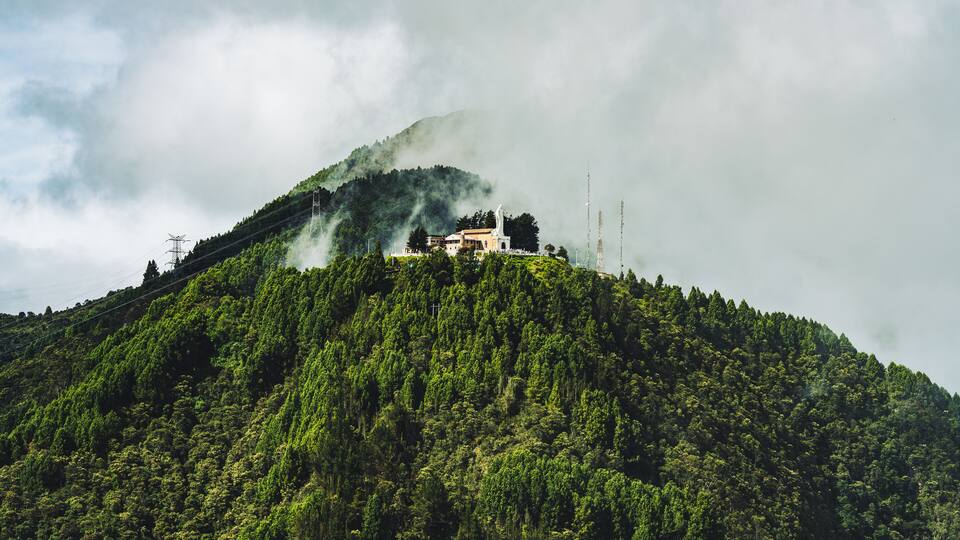 A mountain with a building on top, surrounded by dense green forest and clouds.