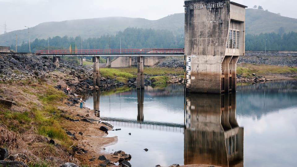 A dam with a bridge crossing over a calm river, surrounded by hills and a rocky shoreline.