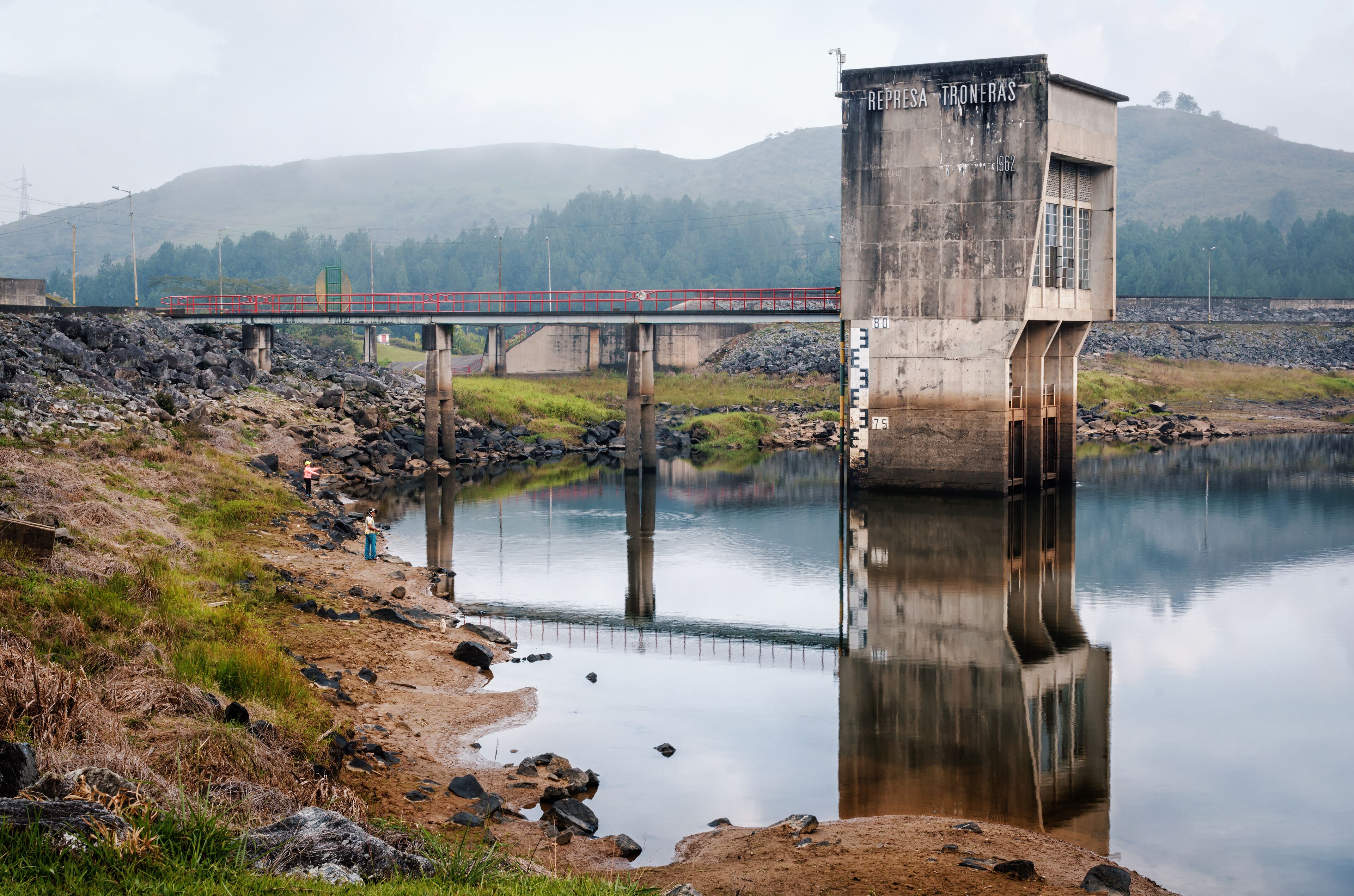 A dam with a bridge crossing over a calm river, surrounded by hills and a rocky shoreline.