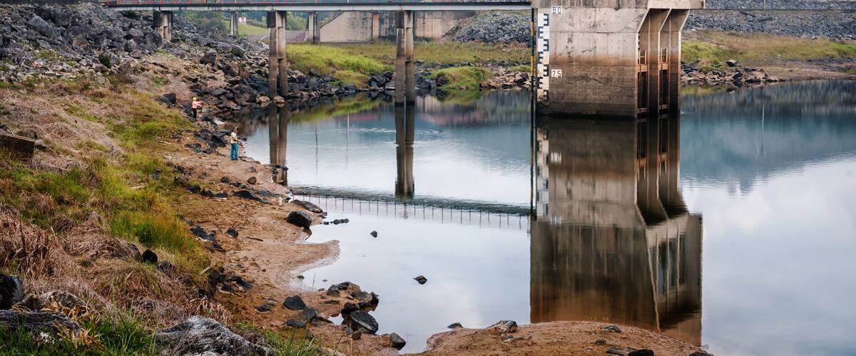 A dam with a bridge crossing over a calm river, surrounded by hills and a rocky shoreline.