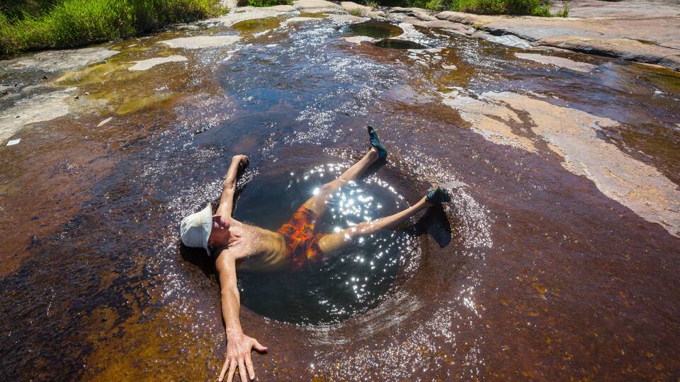 A person lying in a shallow, rocky stream surrounded by greenery.