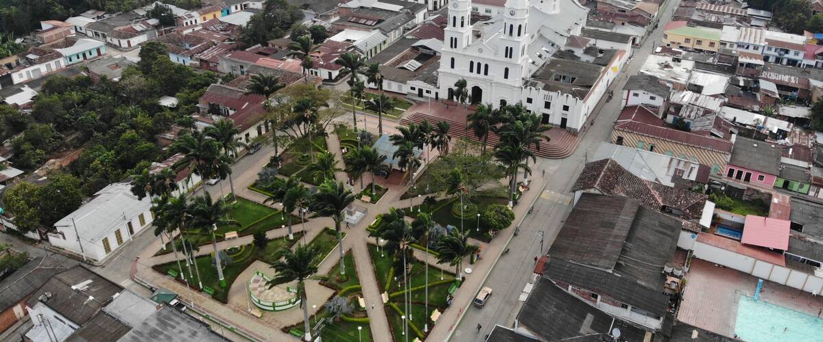 An aerial view of a town with a prominent white church with a dome and a tall bell tower, surrounded by residential buildings and palm trees.