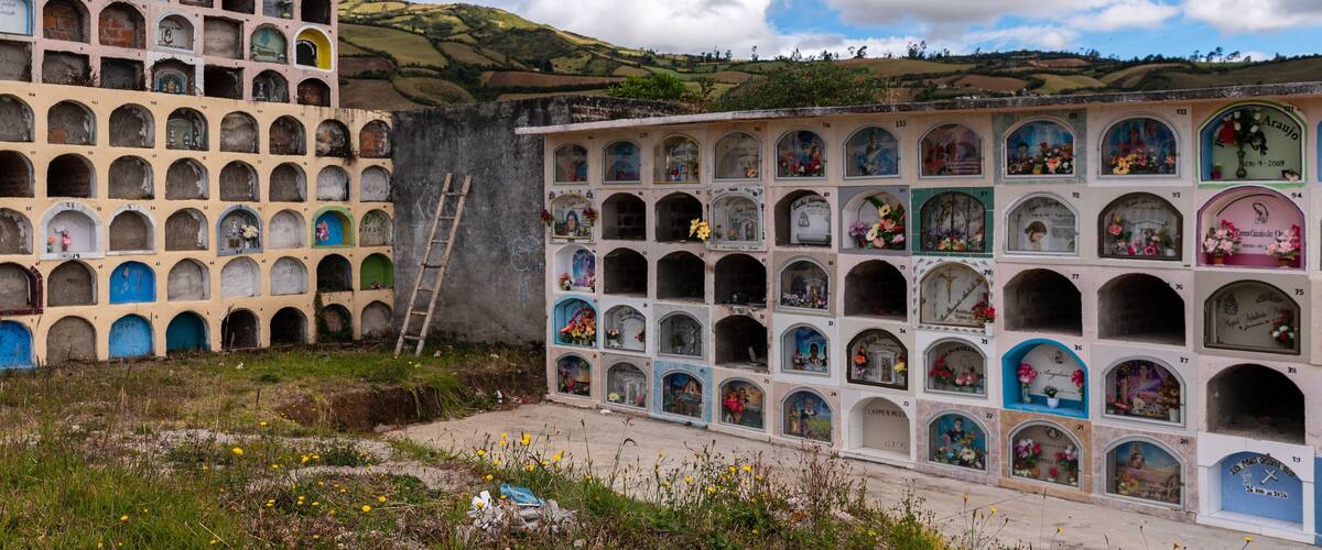 A cemetery with numerous tombs, some adorned with flowers, set against a backdrop of hills and a partly cloudy sky.