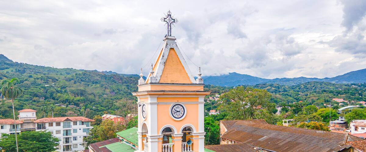 A church with a clock tower, surrounded by residential buildings and lush green hills.