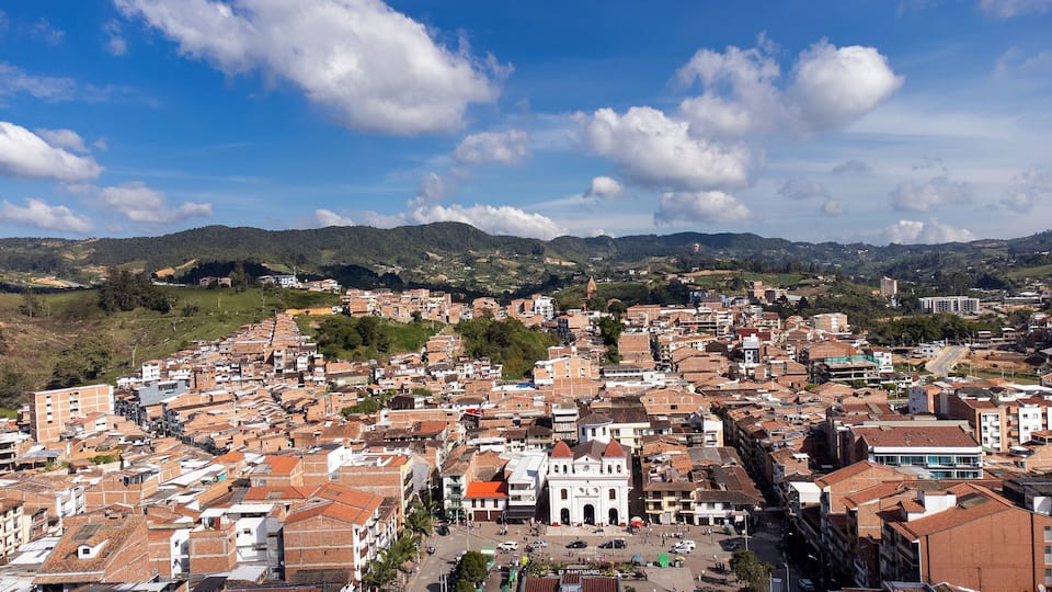 A densely packed urban area with red-tiled rooftops, a prominent white church, and rolling hills in the background.