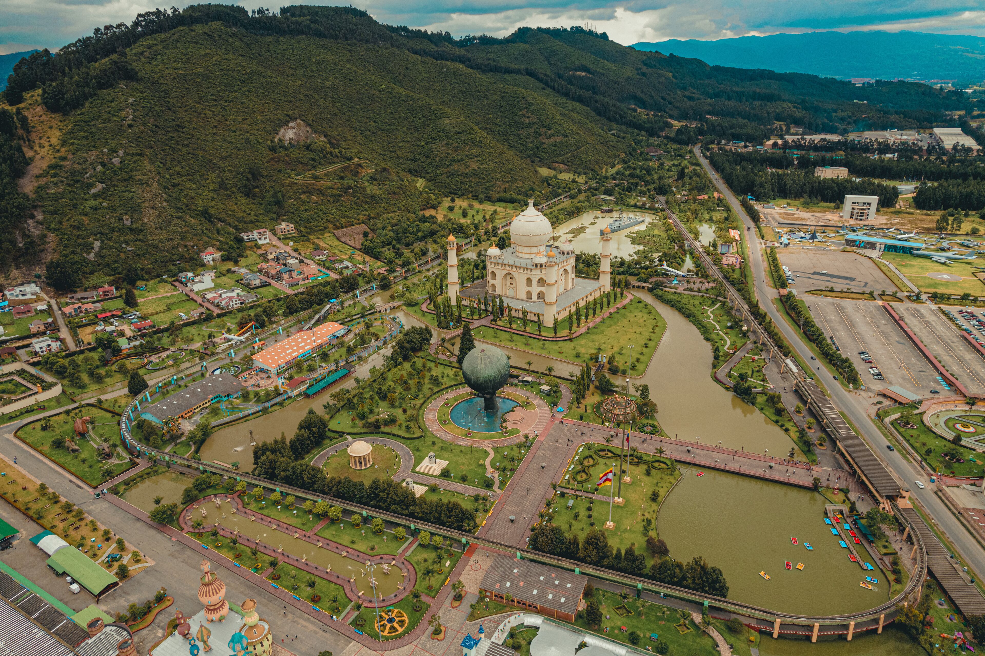 A large mosque with a dome, surrounded by a park with a lake, a bridge, and a mountainous backdrop.