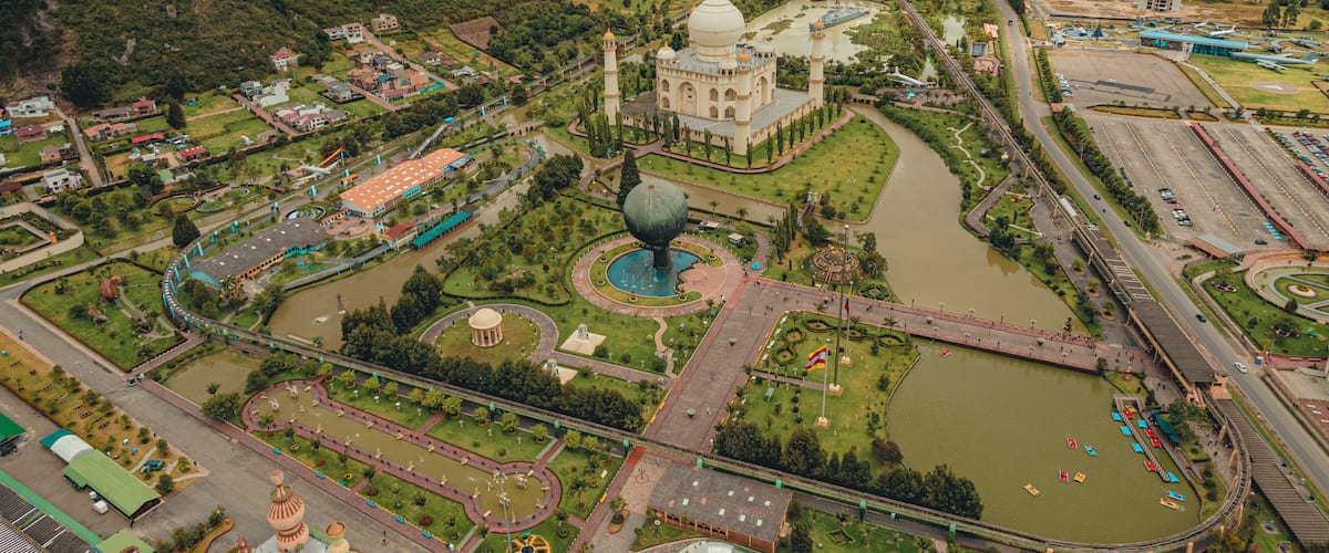 A large mosque with a dome, surrounded by a park with a lake, a bridge, and a mountainous backdrop.