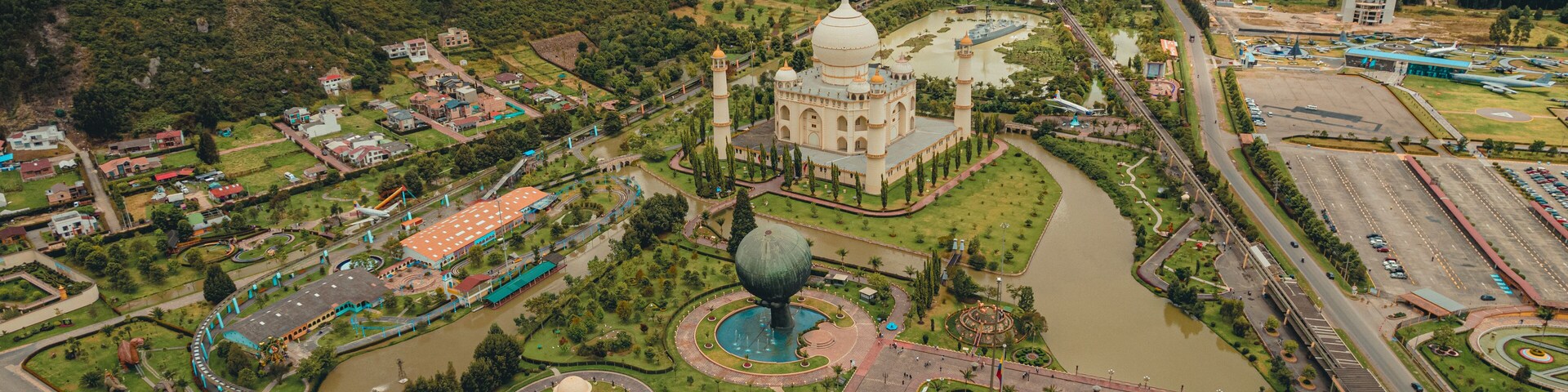 A large mosque with a dome, surrounded by a park with a lake, a bridge, and a mountainous backdrop.