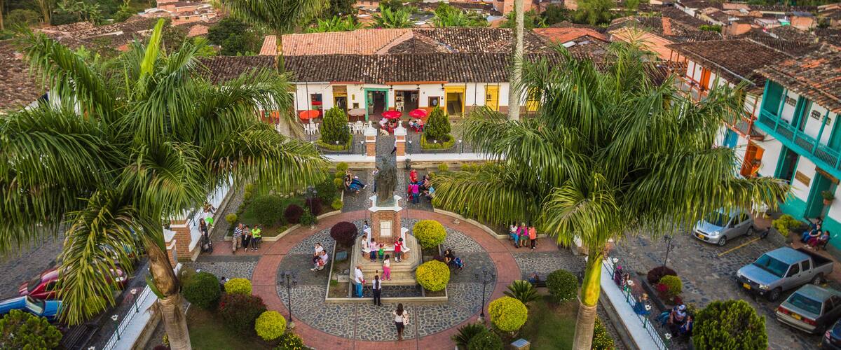 A town square with a central fountain, surrounded by palm trees and buildings with red-tiled roofs.