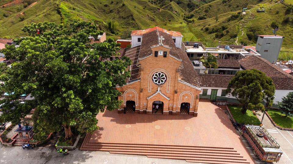 A church with a large circular window and a cross on the roof, surrounded by lush greenery and hills.
