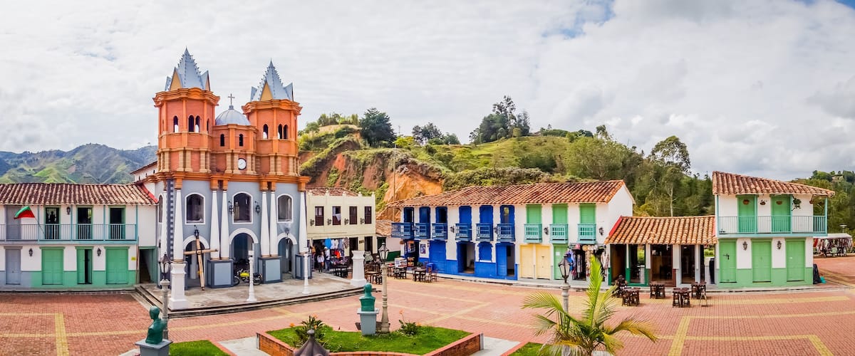 A historic plaza with colorful buildings, a church with two towers, and a mountainous backdrop.