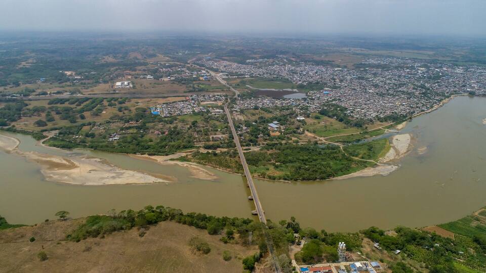 An aerial view of a river with a bridge, a town, and surrounding greenery.
