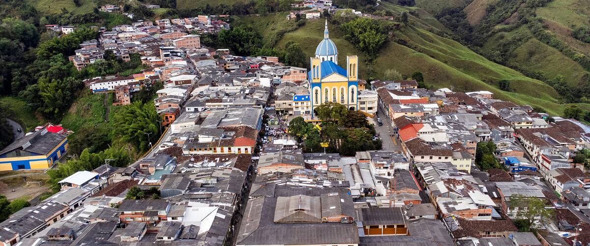 An aerial view of a town with a prominent church with a blue dome and a white spire, surrounded by densely packed buildings and hilly terrain.