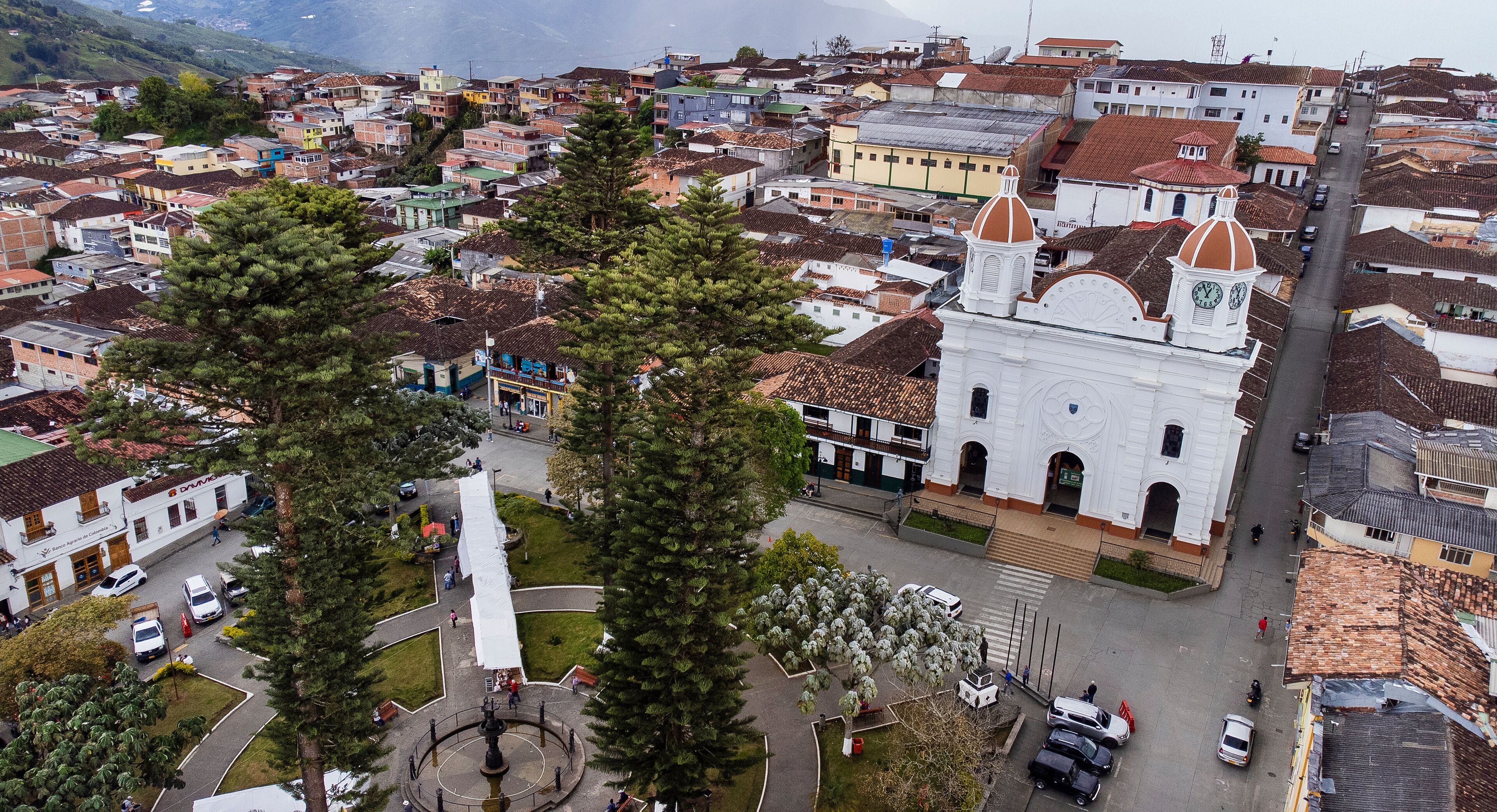 A town with a prominent white church with a dome, surrounded by residential buildings and a central plaza with a fountain.