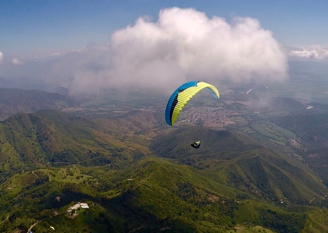 A paraglider flying over a mountainous landscape with lush green hills and a town below.