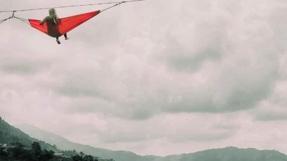 A person hanging from a red fabric between two wires, with a cloudy sky and green hills in the background.