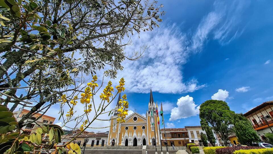 A historic church with a tall bell tower, surrounded by lush greenery and a clear blue sky.
