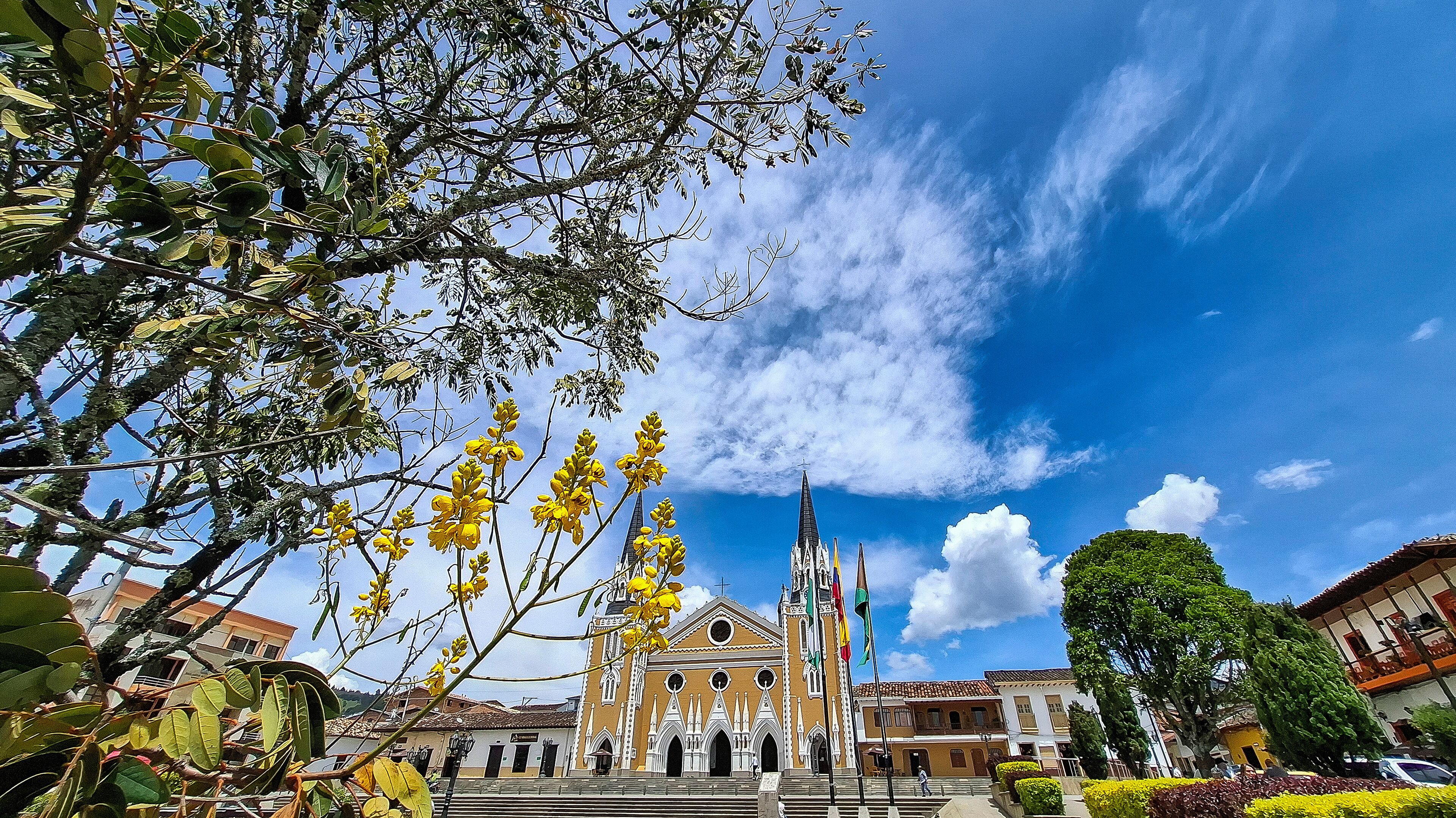 A historic church with a tall bell tower, surrounded by lush greenery and a clear blue sky.
