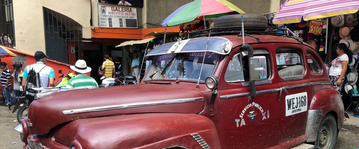 A vintage red car with "Servicio Publico" and "Taxima" on its side parked in a busy street.