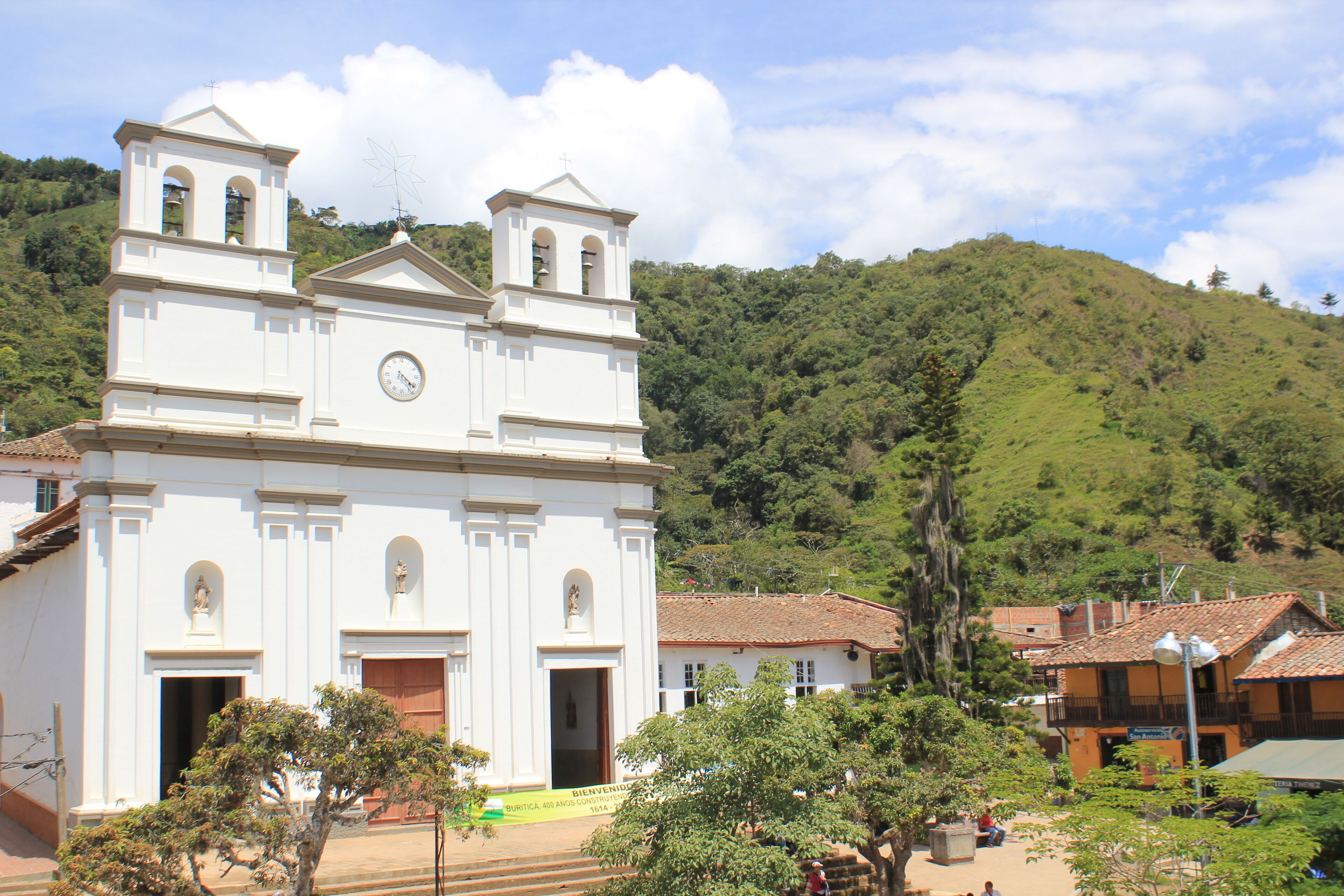 A white church with a bell tower, surrounded by trees and a hillside.