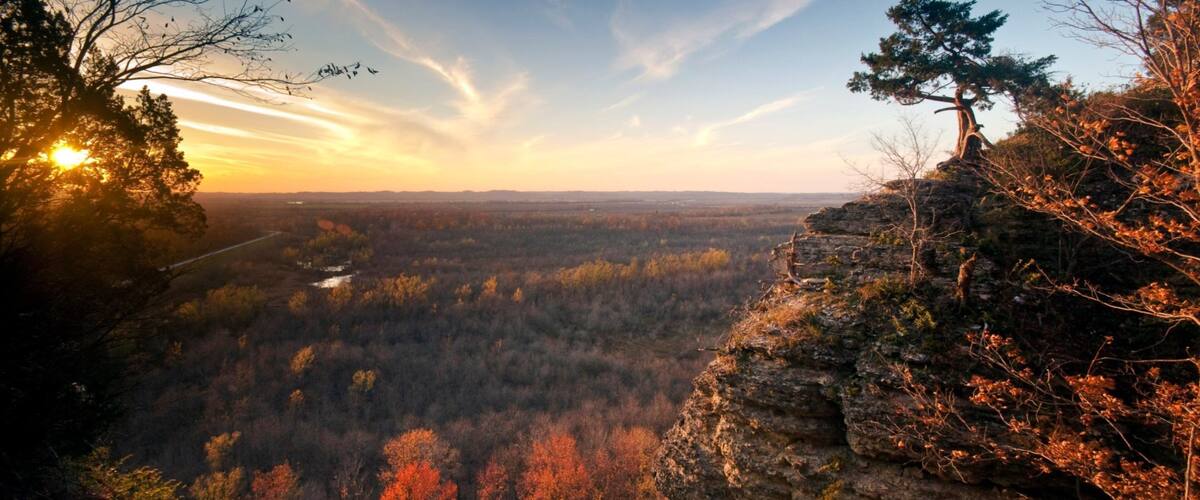 A forested valley with autumn foliage, a rocky cliff, and a sunset.