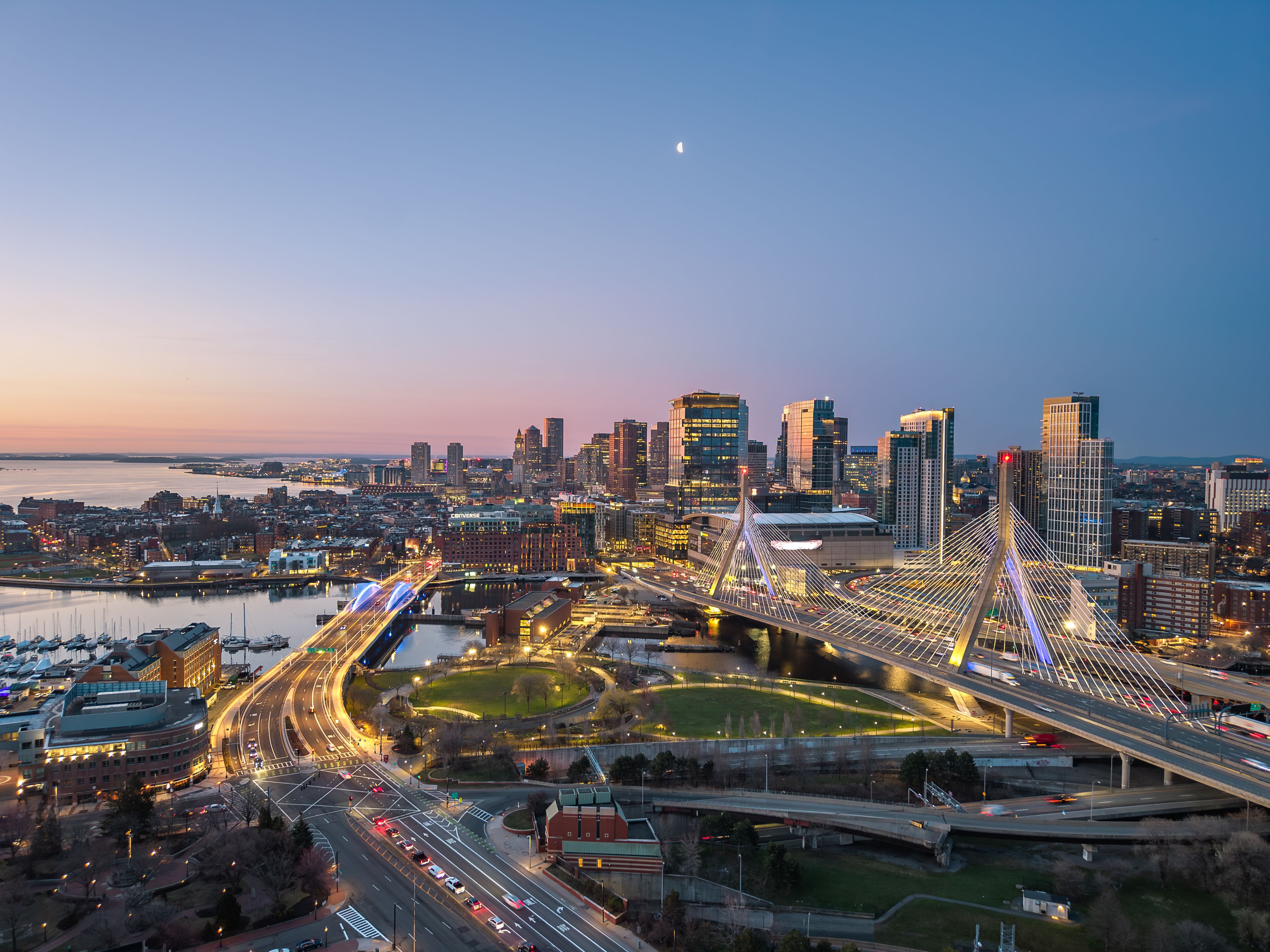A cityscape at dusk with a prominent cable-stayed bridge, high-rise buildings, and a river.