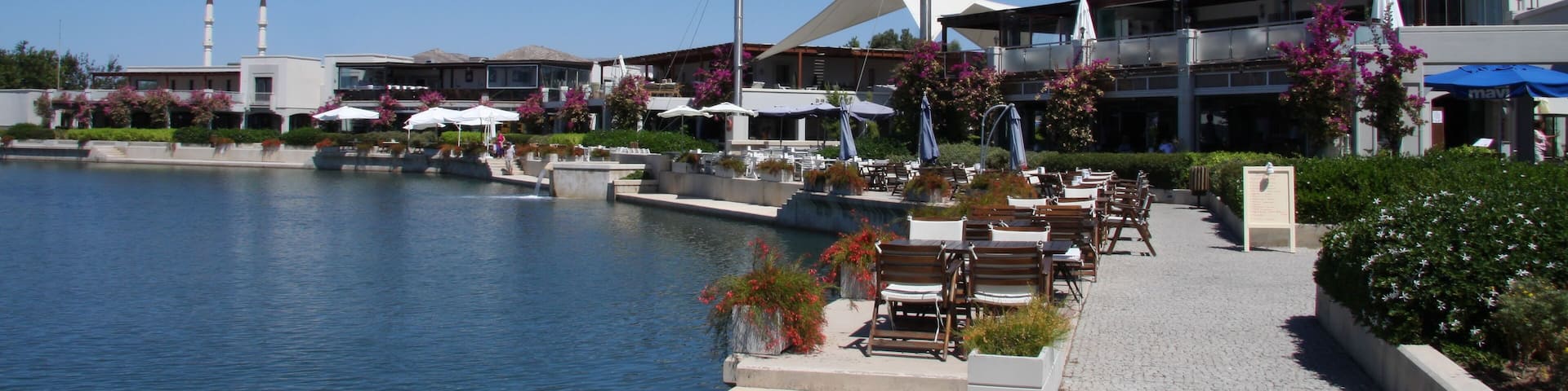 Un paseo marítimo con mesas al aire libre, un edificio moderno con una estructura de techo distintiva y un cielo azul despejado.