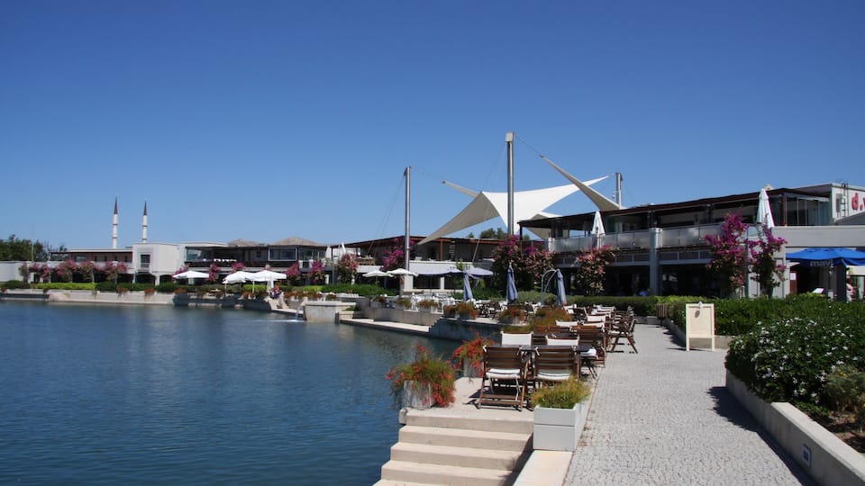 Un paseo marítimo con mesas al aire libre, un edificio moderno con una estructura de techo distintiva y un cielo azul despejado.