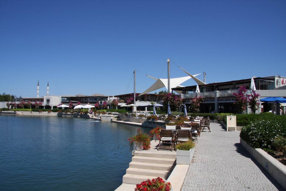 Un paseo marítimo con mesas al aire libre, un edificio moderno con una estructura de techo distintiva y un cielo azul despejado.