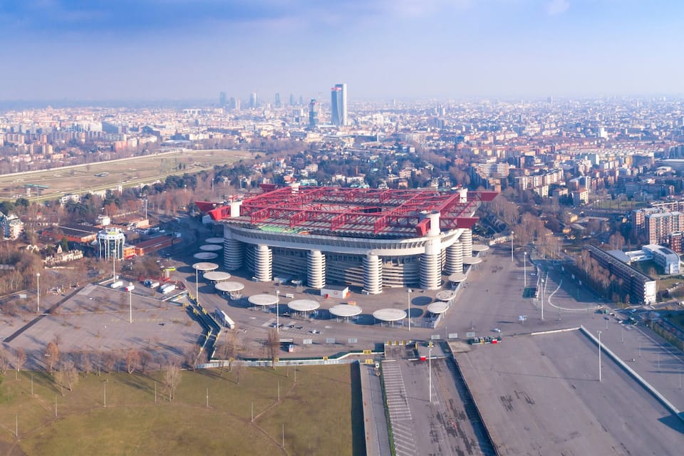 Vista aérea de uma cidade com um grande edifício circular com telhado vermelho, cercado por ruas e edifícios residenciais.