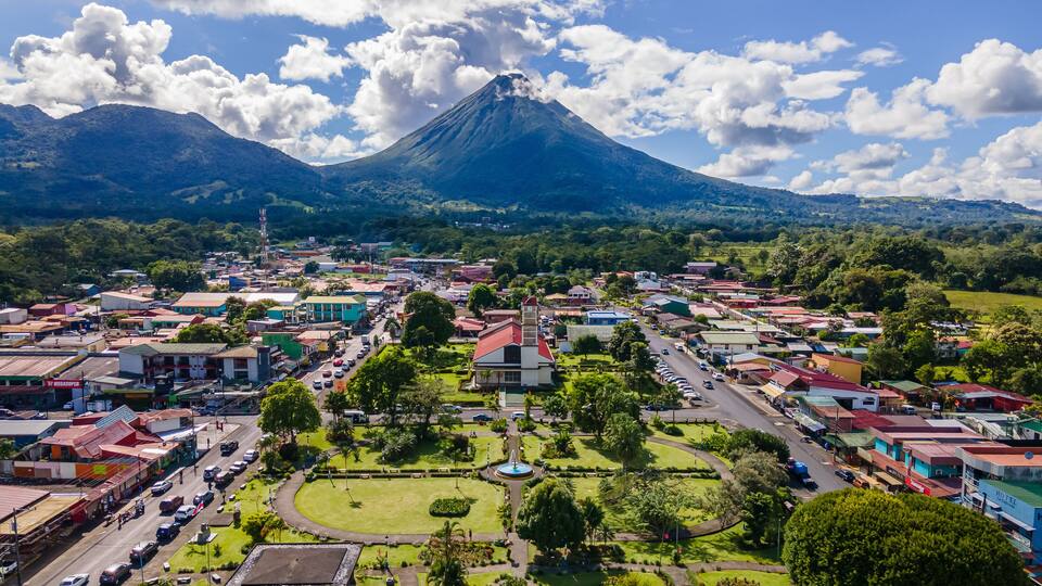 A town with a prominent volcano in the background, surrounded by lush greenery and colorful buildings.