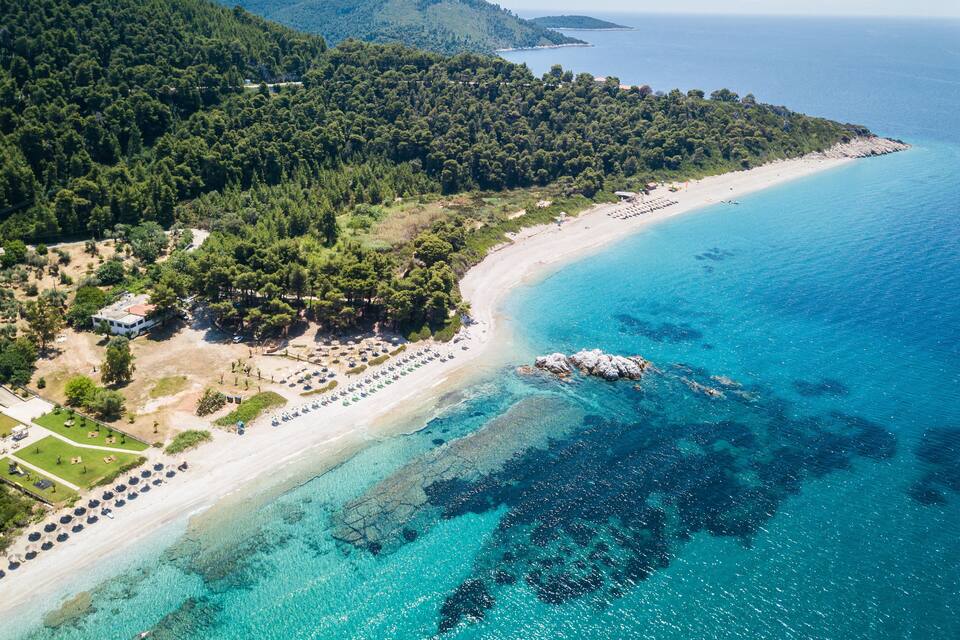 A coastal landscape with a beach, clear turquoise waters, and a forested hillside.