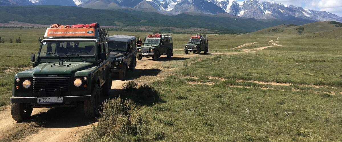 A convoy of military vehicles on a dirt road with snow-capped mountains in the background.