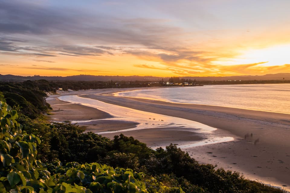 A coastal landscape with a curved shoreline, dense greenery, and a sunset over the water.