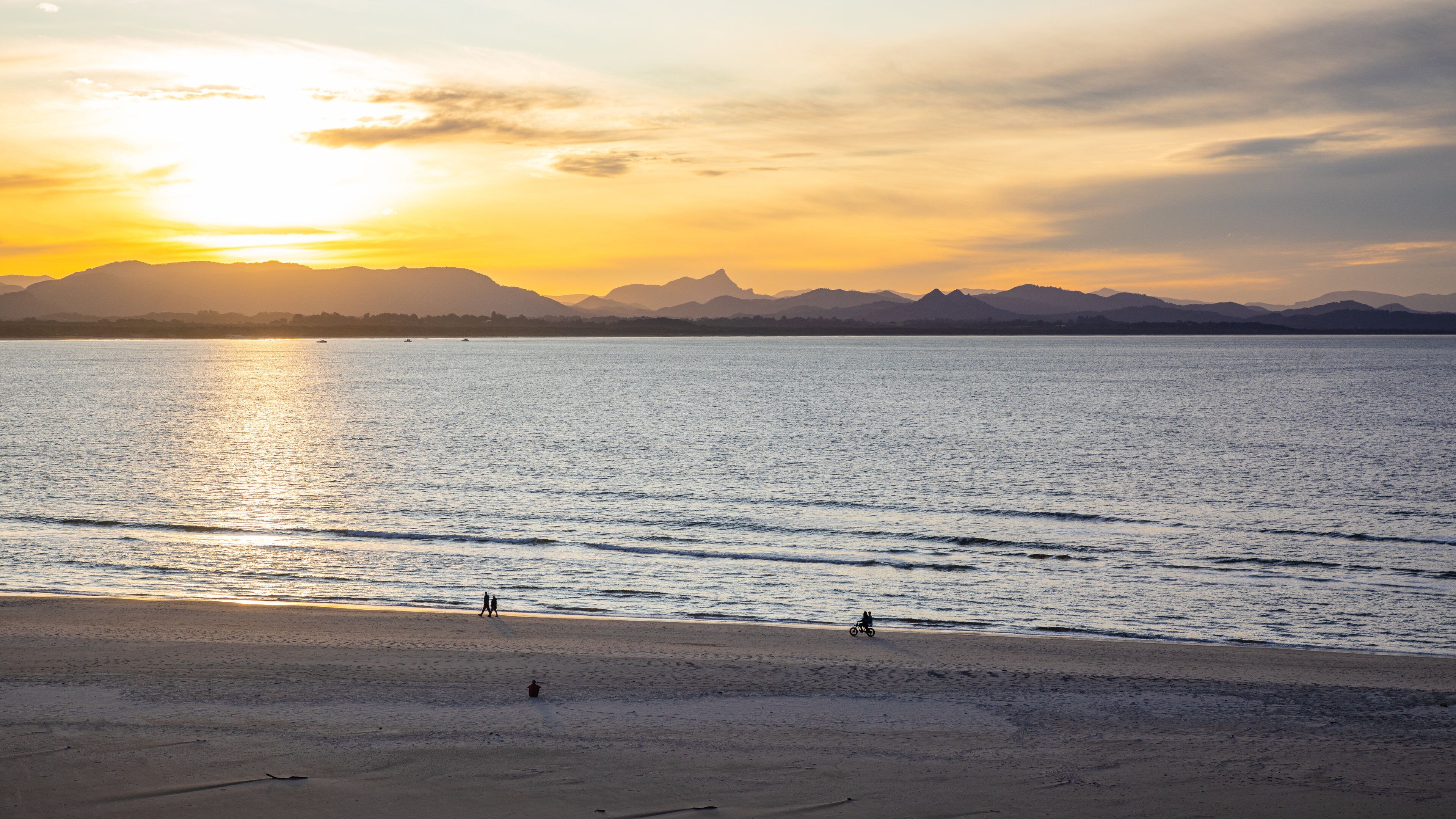 Une plage au coucher du soleil, avec une mer calme et des montagnes à l’horizon.
