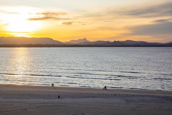 A beach with a sunset, calm sea, and distant mountains.