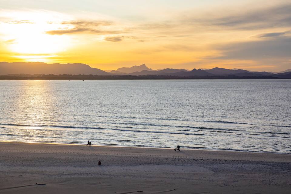 Una playa con un atardecer, mar tranquilo y montañas a lo lejos.