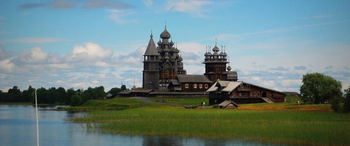 A historic wooden church with multiple domes and a large bell tower by a calm lake.