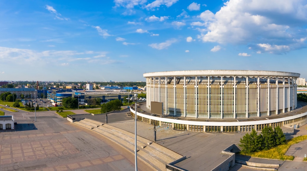 Een groot, rond gebouw met een centrale binnenplaats, omringd door een stadsgezicht met diverse gebouwen en een heldere lucht.
