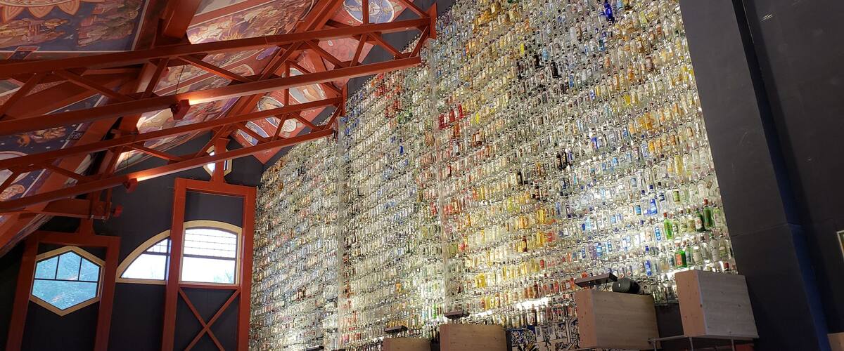 A large collection of bottles arranged on a wall in a room with a high ceiling and wooden beams.