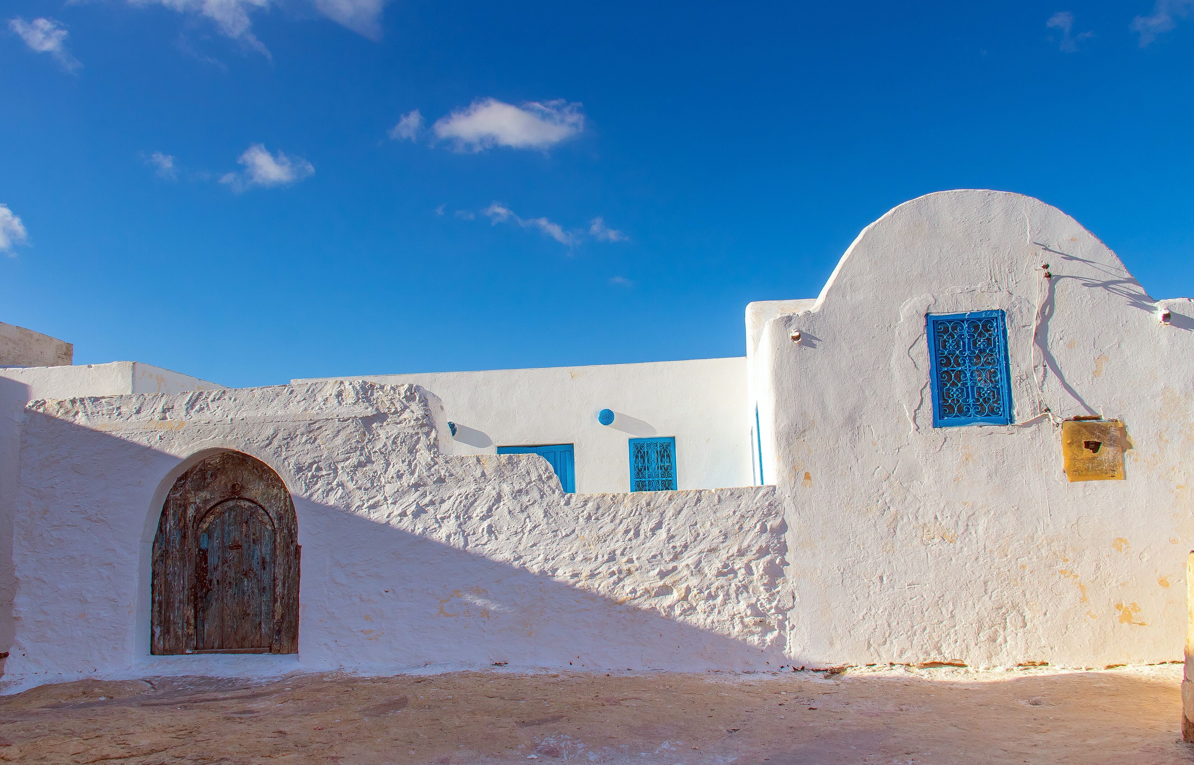 A white building with blue doors and windows, a stone wall, and a clear blue sky.