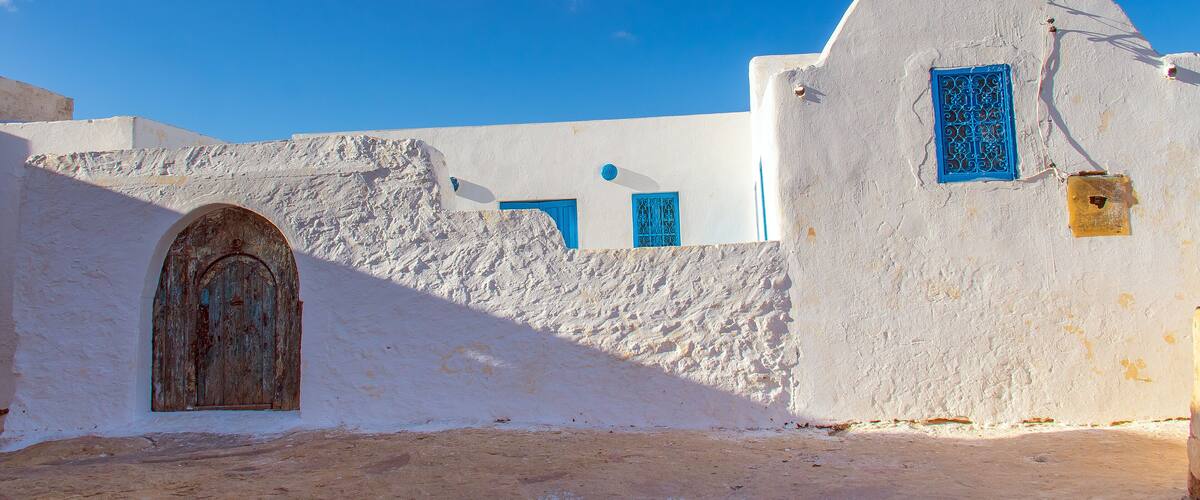 A white building with blue doors and windows, a stone wall, and a clear blue sky.