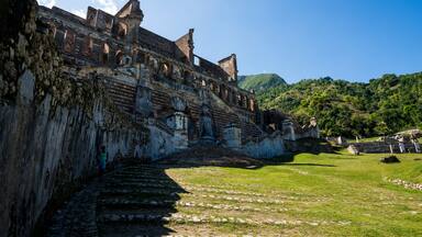 An ancient stone structure with ruins and a staircase leading up to it, surrounded by green hills.
