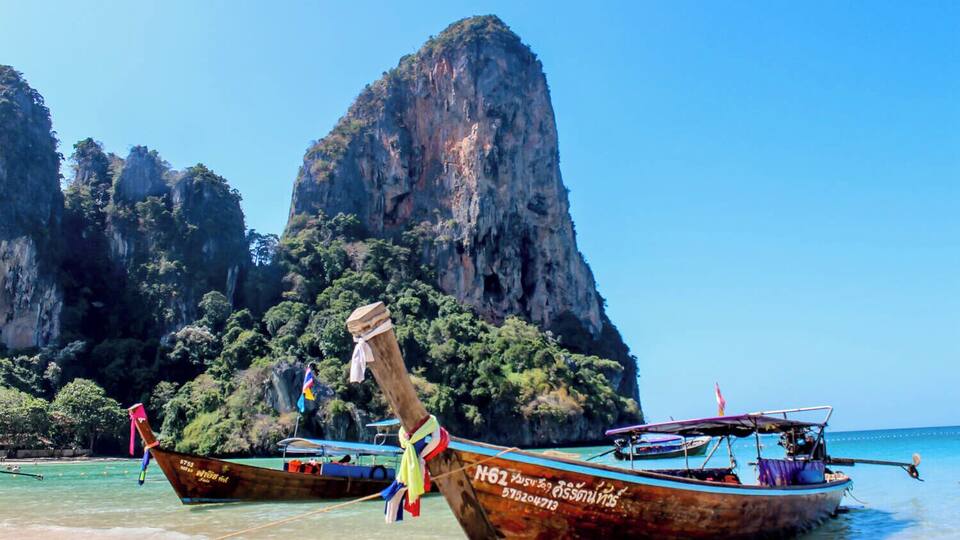 Due imbarcazioni tradizionali ormeggiate su una spiaggia, con una grande formazione rocciosa sullo sfondo.